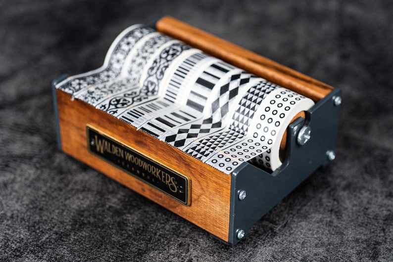 May include: A wooden and metal tape dispenser with six rolls of black and white patterned washi tape. The dispenser has a label that reads "Walden Woodworkers".