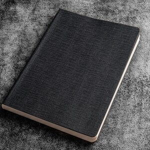K&ouml;nnte beinhalten: Ein schwarzes Notizbuch mit strukturiertem Einband. Das Notizbuch ist geschlossen und liegt flach auf einer grauen, strukturierten Oberfl&auml;che.