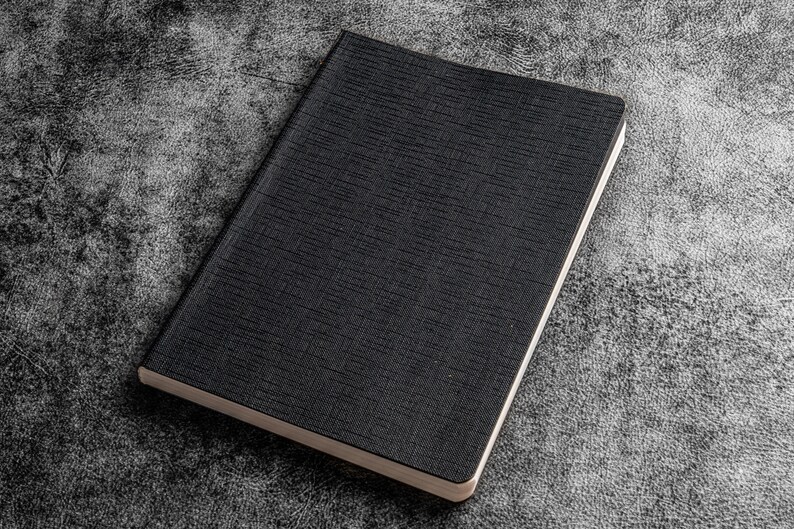 K&ouml;nnte beinhalten: Ein schwarzes Notizbuch mit strukturiertem Einband. Das Notizbuch ist geschlossen und liegt flach auf einer grauen, strukturierten Oberfl&auml;che.