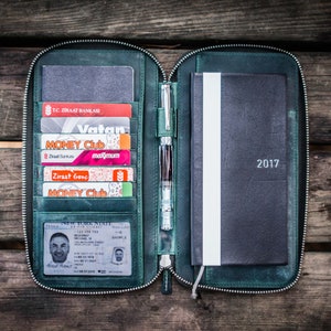 Peut inclure: Un portefeuille en cuir vert zippé avec plusieurs emplacements pour cartes et un porte-stylo. Le portefeuille est ouvert pour révéler un carnet noir avec l'année 2017 imprimée sur la couverture.