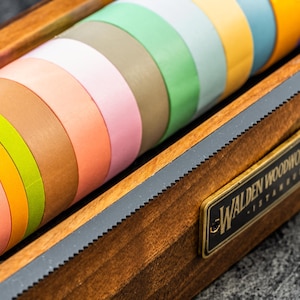 May include: A wooden box with a brass plaque that reads "Walden Woodworkers - Istanbul" holds a collection of colorful washi tapes.