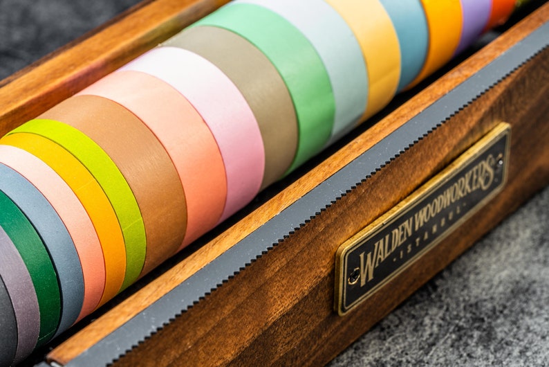 May include: A wooden box with a brass plaque that reads "Walden Woodworkers - Istanbul" holds a collection of colorful washi tapes.