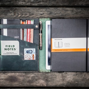 Może przedstawiać: Zielony skórzany pokrowiec na notes z czarnym notesem, długopisem, ołówkami i notesem Field Notes w środku. Pokrowiec ma kieszeń na kartę lub inne małe przedmioty. Pokrowiec na notes jest otwarty, a notes jest widoczny. Notes to Moleskine Classic Collection Ruled Notebook.