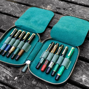 May include: A teal zippered pen case with two rows of slots for holding pens. The case is open and filled with a variety of pens with different colored barrels and gold accents.