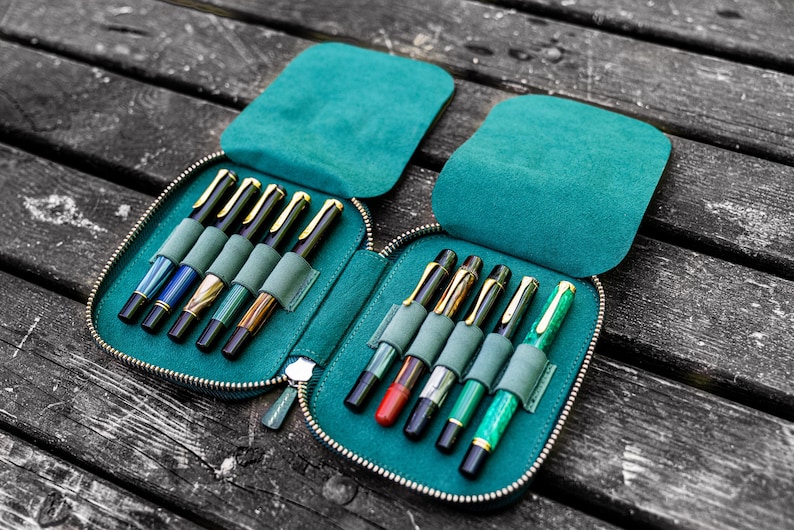 May include: A teal zippered pen case with two rows of slots for holding pens. The case is open and filled with a variety of pens with different colored barrels and gold accents.