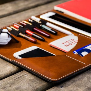 May include: A brown leather portfolio with a variety of pockets and slots for carrying pens, pencils, a phone, a notebook, and cards. The portfolio has white stitching and a black interior.