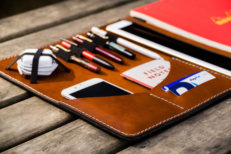 May include: A brown leather portfolio with a variety of pockets and slots for carrying pens, pencils, a phone, a notebook, and cards. The portfolio has white stitching and a black interior.