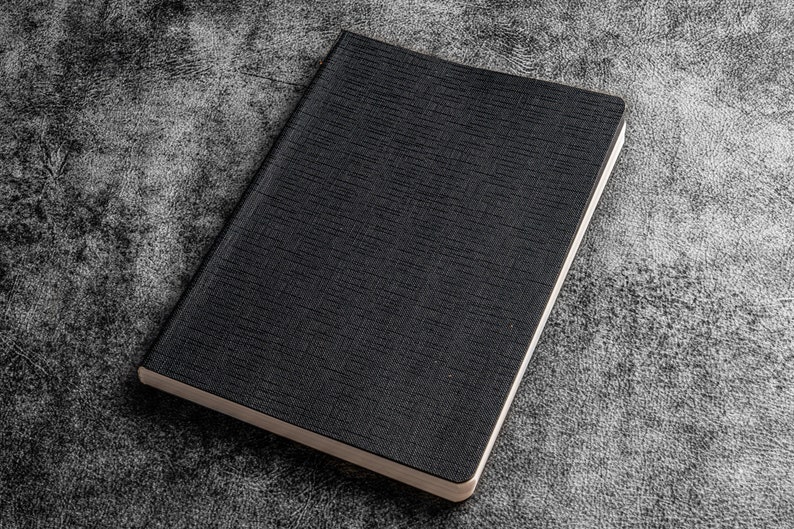 K&ouml;nnte beinhalten: Ein schwarzes Notizbuch mit strukturiertem Einband. Das Notizbuch ist geschlossen und liegt flach auf einer grauen, strukturierten Oberfl&auml;che.