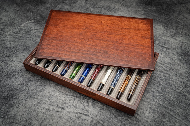 May include: A wooden pen case with a hinged lid, showcasing a collection of colorful fountain pens. The pens feature various designs and colors, including blue, green, and patterned barrels. The case is made of dark wood.