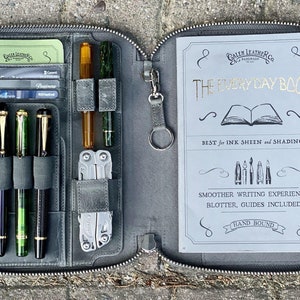 K&ouml;nnte beinhalten: Eine graue Ledertasche mit Rei&szlig;verschluss, die ein Notizbuch mit der Aufschrift "The Everyday Book" enth&auml;lt, sowie den Text "Best for Ink Sheen and Shading" und "Smoother Writing Experience, Blotter, Guides Included, Hand Bound". Die Tasche hat eine Rei&szlig;verschlusstasche mit Stiften, einem Multitool und Karten.