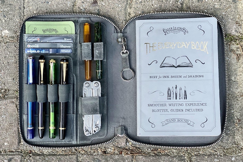 K&ouml;nnte beinhalten: Eine graue Ledertasche mit Rei&szlig;verschluss, die ein Notizbuch mit der Aufschrift "The Everyday Book" enth&auml;lt, sowie den Text "Best for Ink Sheen and Shading" und "Smoother Writing Experience, Blotter, Guides Included, Hand Bound". Die Tasche hat eine Rei&szlig;verschlusstasche mit Stiften, einem Multitool und Karten.