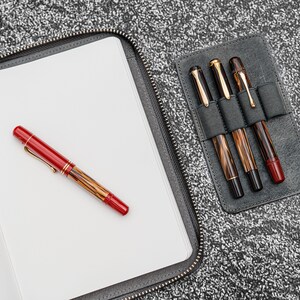 K&ouml;nnte beinhalten: Ein schwarzes Ledernotizbuch mit Rei&szlig;verschluss und Stifthalter. Das Notizbuch ist ge&ouml;ffnet und hat leere wei&szlig;e Seiten. Ein roter und brauner F&uuml;llfederhalter liegt auf den ge&ouml;ffneten Seiten. Ein separater schwarzer Lederstifthalter mit drei braunen und goldenen F&uuml;llfederhaltern ist rechts zu sehen.