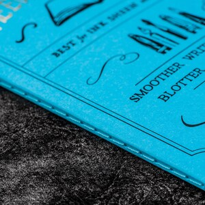 Pu&ograve; includere: Un quaderno blu con il testo "The Ever After" e "Best for Ink & Quill" sulla copertina. Il quaderno ha anche il testo "Smoother Writing Experience, Blotter, Guides Included, Hand Crafted" sulla copertina.