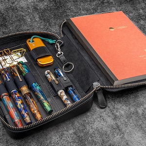 May include: A black leather zippered case with a notebook, pens, and a small multi-tool. The case has a zipper closure and a variety of pockets and compartments for organizing items.