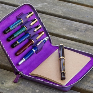 May include: A purple leather pen case with a zipper closure, containing six pens and a small notebook. The case is open and the pens are visible.