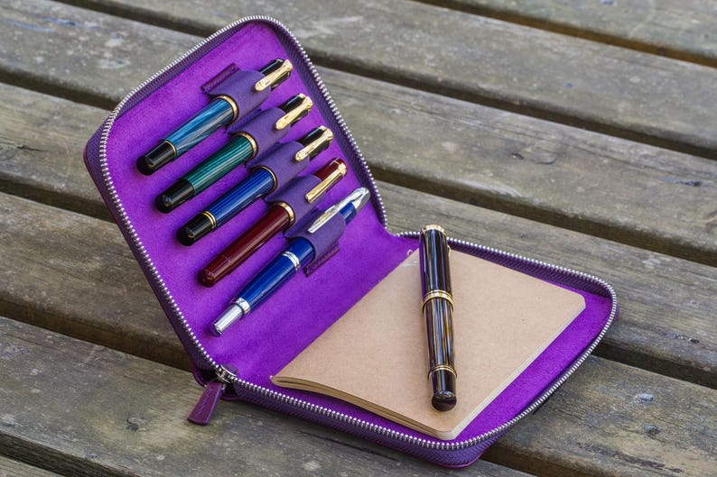 May include: A purple leather pen case with a zipper closure, containing six pens and a small notebook. The case is open and the pens are visible.