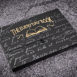 K&ouml;nnte beinhalten: Ein schwarzes Buch mit goldener Beschriftung "THE EVERY DAY BOOK" und einer goldenen Illustration eines ge&ouml;ffneten Buches auf dem Einband. Das Buch ist von handschriftlichem Text umgeben.