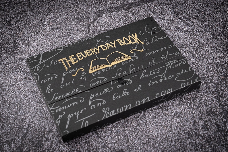 K&ouml;nnte beinhalten: Ein schwarzes Buch mit goldener Beschriftung "THE EVERY DAY BOOK" und einer goldenen Illustration eines ge&ouml;ffneten Buches auf dem Einband. Das Buch ist von handschriftlichem Text umgeben.