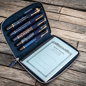 Op de afbeelding: Een blauwe leren ritshoes met zes pennen vakken en een klein notitieboekje met de tekst "Note Book" op de omslag. Het notitieboekje heeft een blauw-wit geruit patroon.
