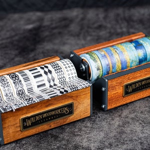 May include: Two wooden tape dispensers with metal accents. The dispensers are filled with colourful and patterned washi tapes. The dispensers have a brass plaque with the text "Walden Woodworkers Istanbul" on them.