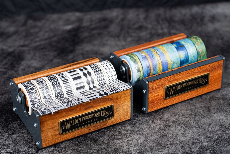 May include: Two wooden tape dispensers with metal accents. The dispensers are filled with colourful and patterned washi tapes. The dispensers have a brass plaque with the text "Walden Woodworkers Istanbul" on them.