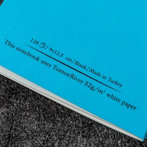Pu&ograve; includere: Un quaderno blu con il testo "This notebook uses Tomoe River 52g/m&sup2; white paper 128/9x12.5 cm/Blank/Made in Turkey" stampato sulla copertina posteriore.