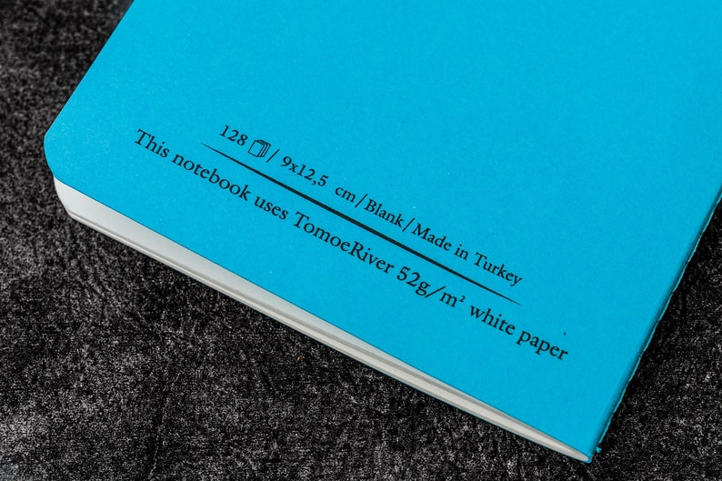 Pu&ograve; includere: Un quaderno blu con il testo "This notebook uses Tomoe River 52g/m&sup2; white paper 128/9x12.5 cm/Blank/Made in Turkey" stampato sulla copertina posteriore.