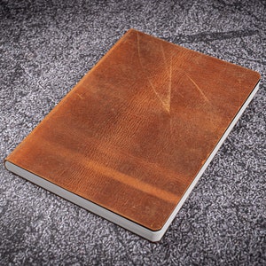 May include: A rectangular brown leather journal with a white-edged page block. The cover has a textured appearance with visible grain and natural markings. The notebook is closed and lies flat on a textured grey surface.