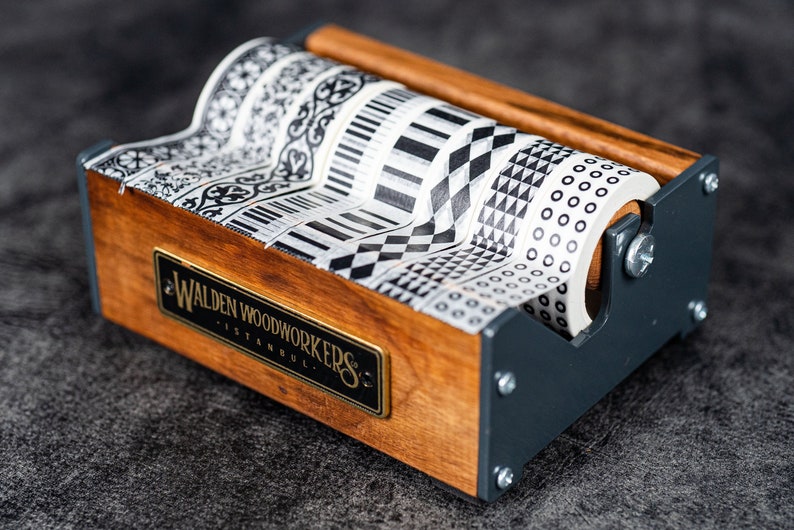 May include: A wooden tape dispenser with a black metal frame holds five rolls of black and white patterned washi tape. The dispenser has a brass plaque that reads "Walden Woodworkers Istanbul."
