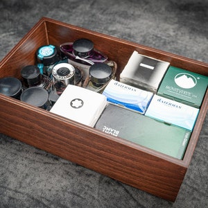 Puede incluir: Una caja de madera llena de varios artículos de papelería. La caja contiene frascos de tinta, cartuchos de tinta y una tarjeta blanca con un emblema negro. La caja es de madera marrón oscuro y está sobre una superficie gris.