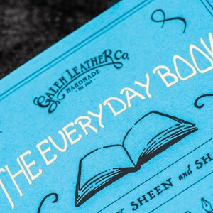 Pu&ograve; includere: Un quaderno blu con il testo "The Everyday Book" in lettere bianche. Il quaderno &egrave; di Galen Leather Co. e presenta un'illustrazione in bianco e nero di un libro aperto.