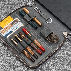 K&ouml;nnte beinhalten: Eine graue Leder-Rei&szlig;verschlusstasche mit einem schwarzen Notizbuch, mehreren Stiften und einem Schl&uuml;sselanh&auml;nger. Die Tasche hat F&auml;cher f&uuml;r Stifte und Karten. Die Tasche ist ge&ouml;ffnet und der Inhalt ist sichtbar.
