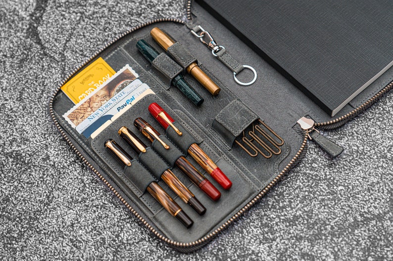 K&ouml;nnte beinhalten: Eine graue Leder-Rei&szlig;verschlusstasche mit einem schwarzen Notizbuch, mehreren Stiften und einem Schl&uuml;sselanh&auml;nger. Die Tasche hat F&auml;cher f&uuml;r Stifte und Karten. Die Tasche ist ge&ouml;ffnet und der Inhalt ist sichtbar.