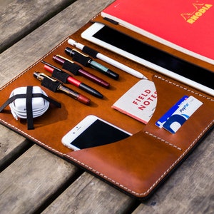 May include: A brown leather portfolio with a variety of pockets and slots for carrying a tablet, phone, pens, and cards. The portfolio is open and laid flat on a wooden surface. The text "FIELD NOTES" is visible on a small notebook inside the portfolio.