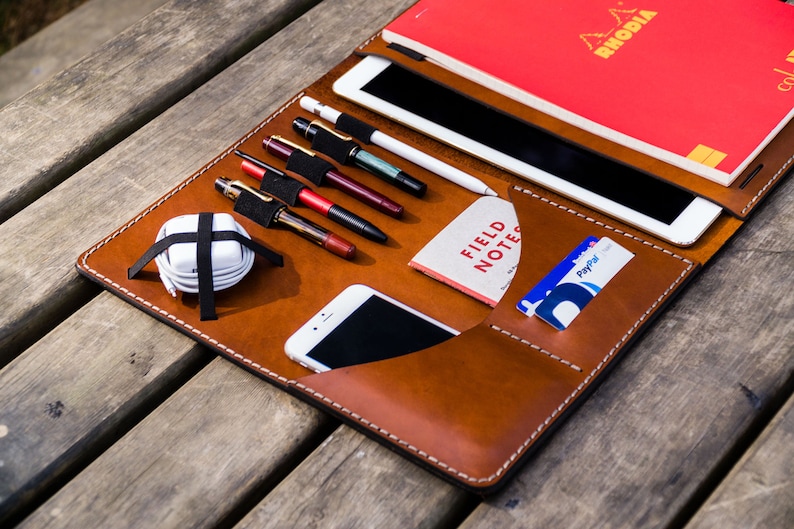 May include: A brown leather portfolio with a variety of pockets and slots for carrying a tablet, phone, pens, and cards. The portfolio is open and laid flat on a wooden surface. The text "FIELD NOTES" is visible on a small notebook inside the portfolio.