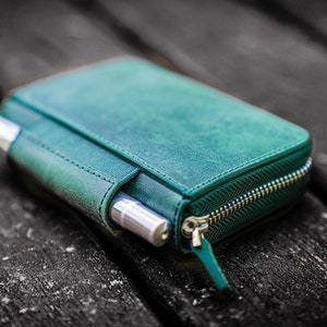 May include: A teal leather zipped pouch with two silver pens inside. The pouch is lying on a dark wooden surface.