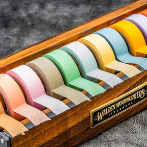 May include: A wooden tape dispenser with a label that reads "Walden Woodworkers - Istanbul" holds a variety of colorful washi tapes. The tapes are in shades of green, blue, yellow, orange, pink, and brown.