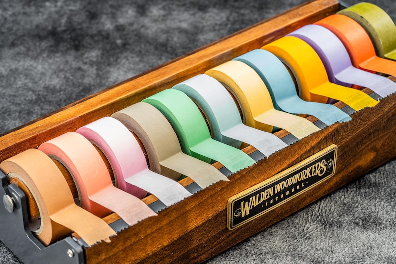 May include: A wooden tape dispenser with a label that reads "Walden Woodworkers - Istanbul" holds a variety of colorful washi tapes. The tapes are in shades of green, blue, yellow, orange, pink, and brown.