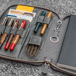 K&ouml;nnte beinhalten: Eine graue Leder-Rei&szlig;verschlusstasche mit einem Notizbuch und Stiften im Inneren. Die Tasche hat mehrere F&auml;cher f&uuml;r Stifte und einen Schl&uuml;sselring. Das Notizbuch ist schwarz mit einem strukturierten Einband.