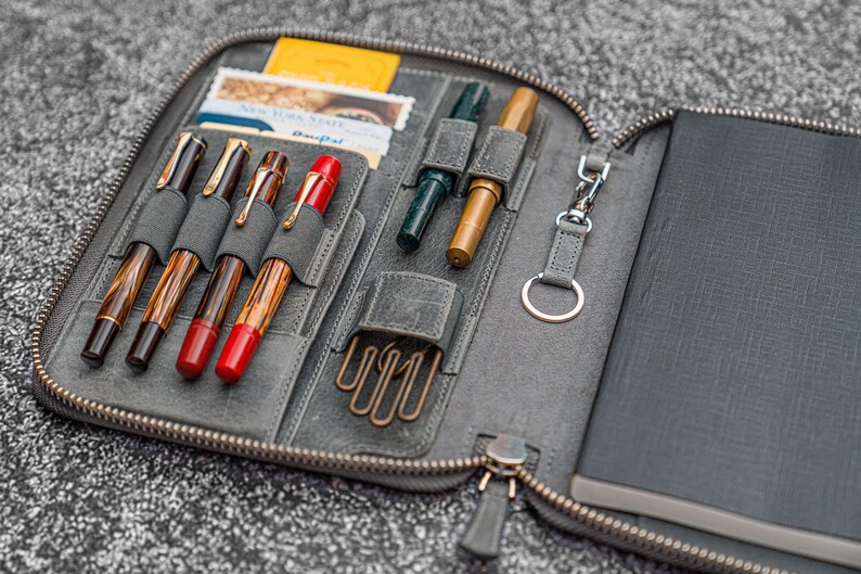 K&ouml;nnte beinhalten: Eine graue Leder-Rei&szlig;verschlusstasche mit einem Notizbuch und Stiften im Inneren. Die Tasche hat mehrere F&auml;cher f&uuml;r Stifte und einen Schl&uuml;sselring. Das Notizbuch ist schwarz mit einem strukturierten Einband.