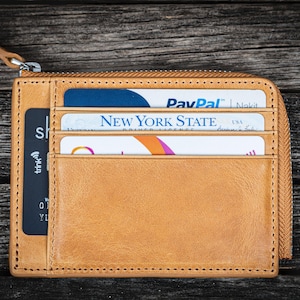 May include: A tan leather zippered wallet with multiple card slots. The wallet is open and contains a black credit card, a New York State driver's license, and a PayPal credit card.