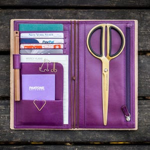 Può includere: Un diario in pelle viola con un paio di forbici color oro, carte di credito e una matita. Il diario è aperto, rivelando tasche e uno scomparto con cerniera. Le forbici hanno manici neri e il testo "BEAUTY TOOLS" sulla lama.