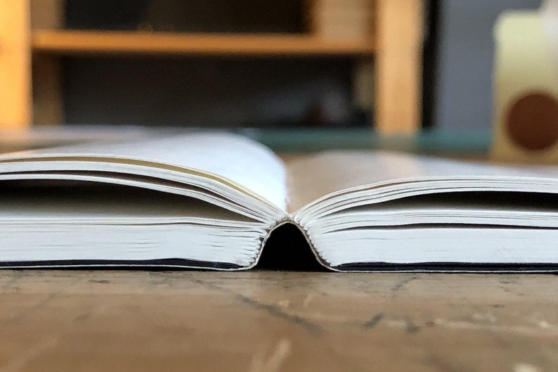 K&ouml;nnte beinhalten: Nahaufnahme eines leeren, wei&szlig;en Notizbuchs mit schwarzem Einband und goldenen Akzenten. Das Notizbuch ist auf einer leeren Seite ge&ouml;ffnet.