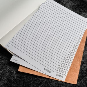K&ouml;nnte beinhalten: Zwei Bl&auml;tter wei&szlig;es Papier mit liniertem und kariert Papierdesign. Das Papier liegt auf einem braunen Ledernotizbuch.