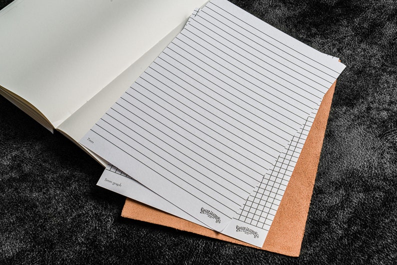 K&ouml;nnte beinhalten: Zwei Bl&auml;tter wei&szlig;es Papier mit liniertem und kariert Papierdesign. Das Papier liegt auf einem braunen Ledernotizbuch.