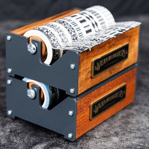 May include: A wooden and metal tape dispenser with two rolls of patterned washi tape. The dispenser has a black metal frame and wooden sides with a brass plaque that reads "Walden Woodworkers - Est. 1978".
