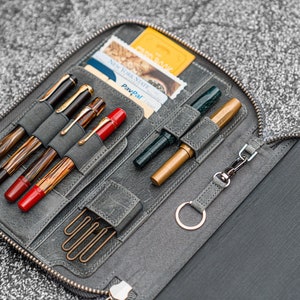 K&ouml;nnte beinhalten: Eine graue Leder-Rei&szlig;verschlusstasche mit mehreren F&auml;chern f&uuml;r Stifte und einem kleinen Fach f&uuml;r Karten. Die Tasche ist ge&ouml;ffnet und zeigt eine Vielzahl von Stiften, darunter einen gr&uuml;nen Stift, einen goldenen Stift und mehrere schwarze und braune Stifte. Es befinden sich auch mehrere B&uuml;roklammern in der Tasche.