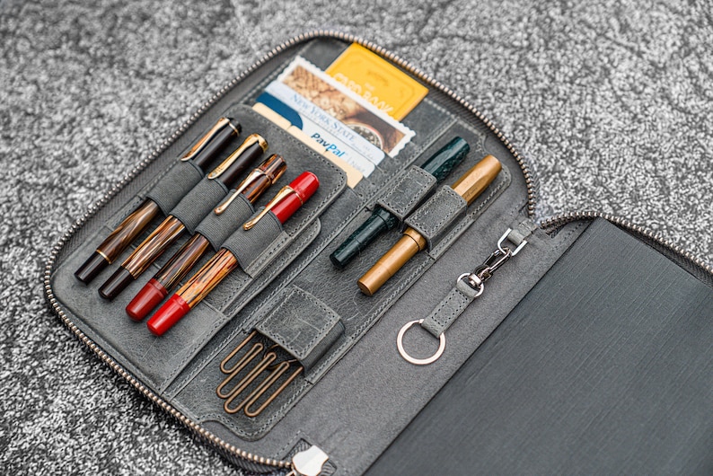 K&ouml;nnte beinhalten: Eine graue Leder-Rei&szlig;verschlusstasche mit mehreren F&auml;chern f&uuml;r Stifte und einem kleinen Fach f&uuml;r Karten. Die Tasche ist ge&ouml;ffnet und zeigt eine Vielzahl von Stiften, darunter einen gr&uuml;nen Stift, einen goldenen Stift und mehrere schwarze und braune Stifte. Es befinden sich auch mehrere B&uuml;roklammern in der Tasche.