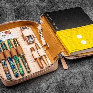 May include: A light brown leather zippered case with a black notebook and several pens inside. The notebook is a Stalogy 365 Days Notebook. The case has a zippered closure and a small pocket on the outside.