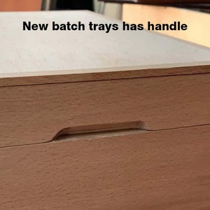 May include: Close-up of a light-colored wooden box with a recessed handle. The box has a smooth, natural wood finish and a simple, rectangular design. The text "New batch trays has handle" is visible.